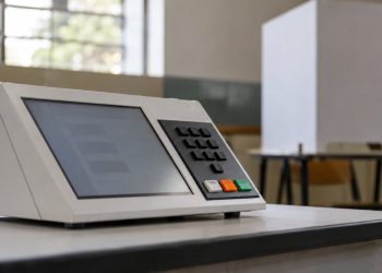 Urna eletrônica em seção eleitoral, com cabine de votação ao fundo