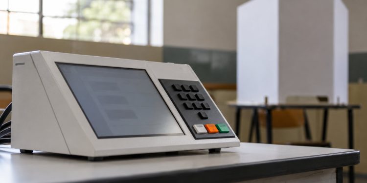 Urna eletrônica em seção eleitoral, com cabine de votação ao fundo